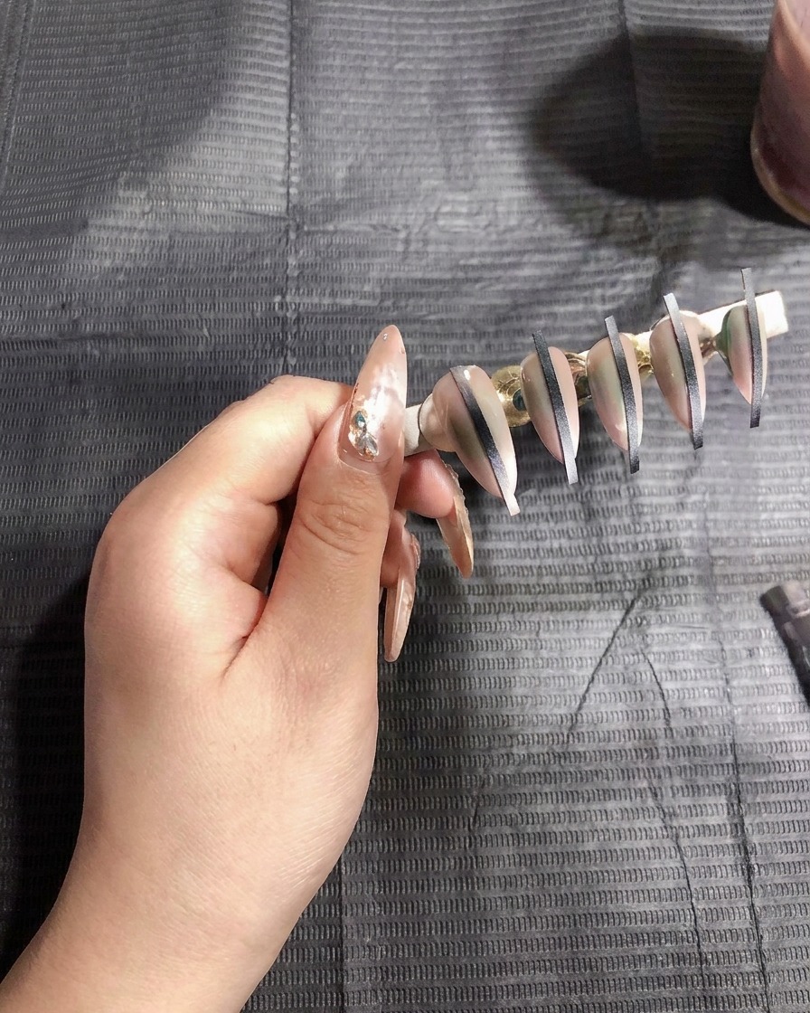 An airbrush gun spraying a thin layer of white gel onto artificial nails mounted on a stand.