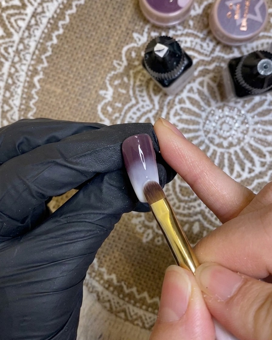 A close-up of a brush applying a dark, rich purple gel onto the tip of a manicured fingernail.