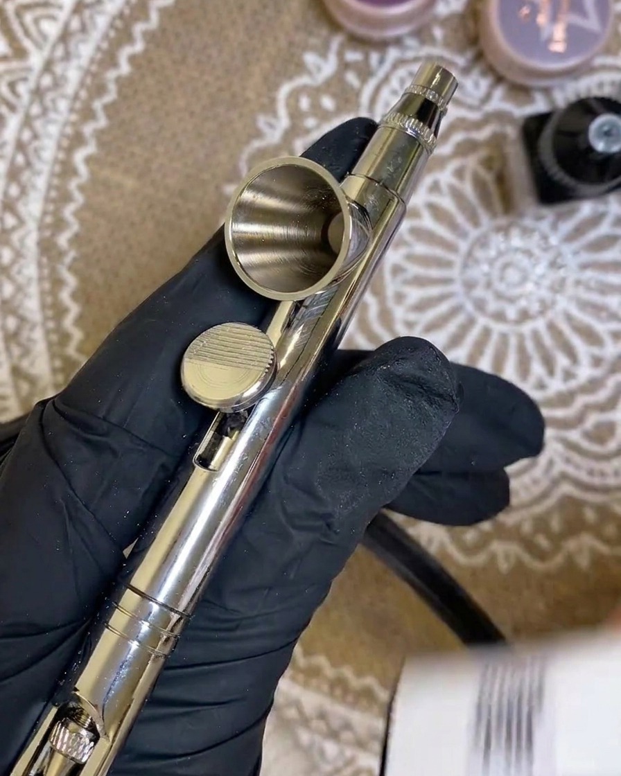 A close-up showing a bottle of white gel being poured into the small metal cup of an airbrush tool.