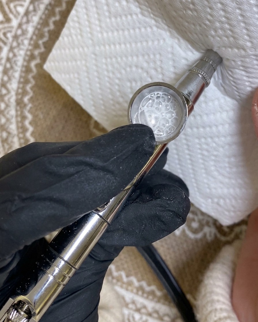 An airbrush tool pointed at a paper towel, showing a test spray pattern of white paint.