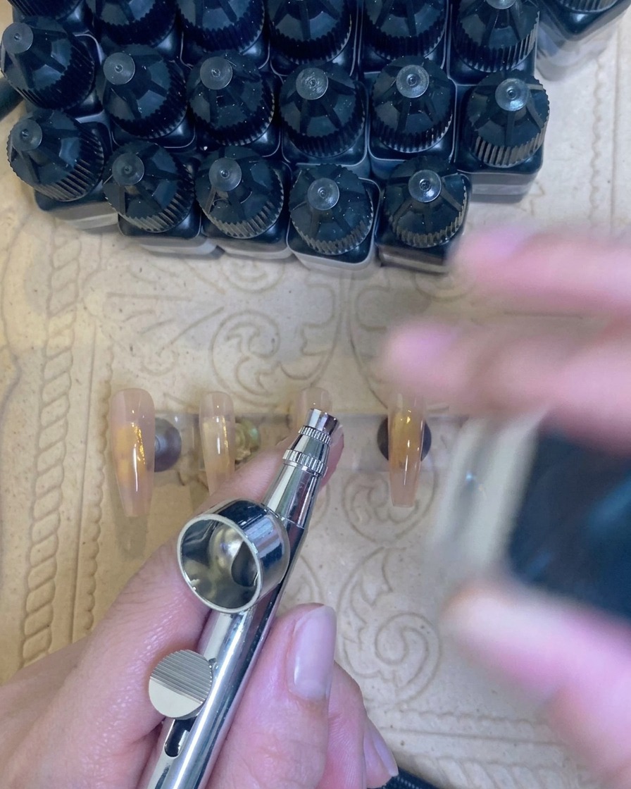 An airbrush tool applying a soft white mist to the tip of a nude nail extension.