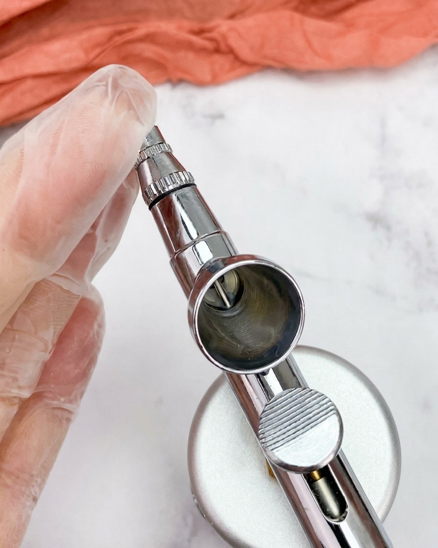 A close-up view of a clean, empty metal airbrush cup attached to the airbrush gun.