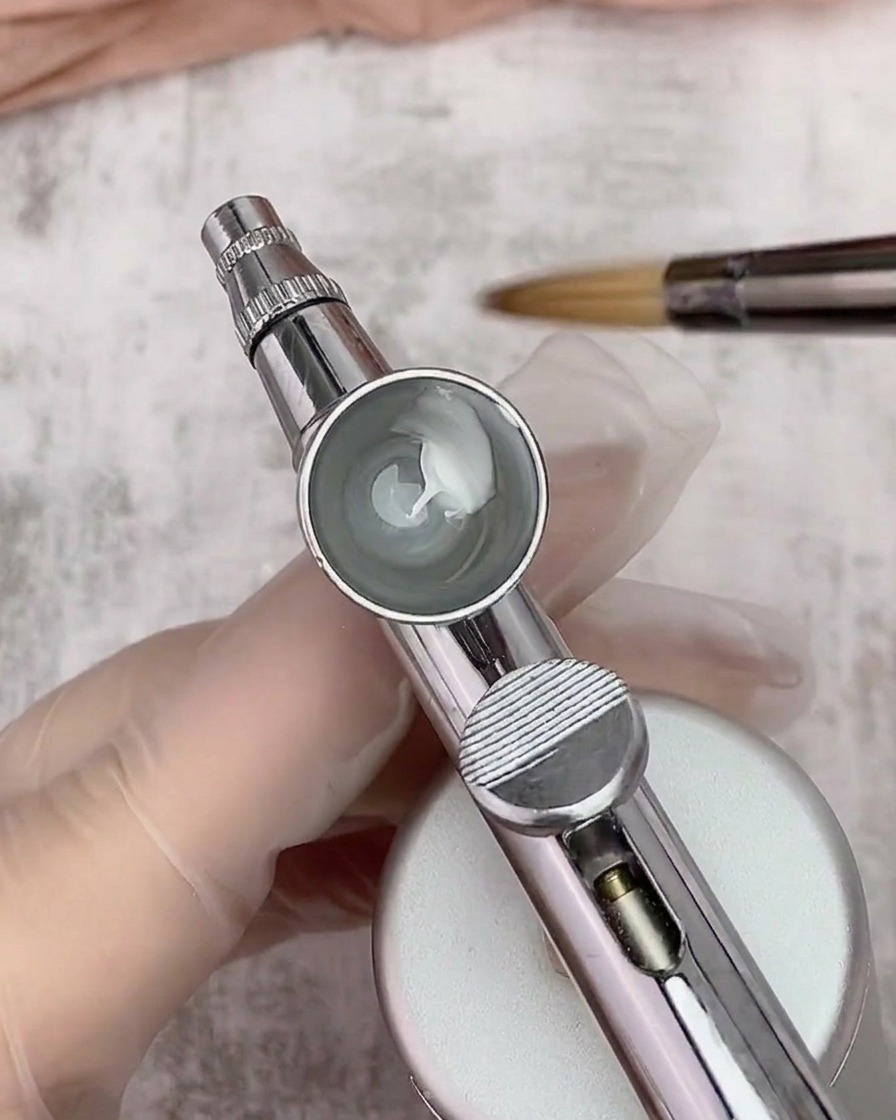 A close-up looking into an airbrush cup containing a mix of white gel polish and thinner, with a brush hovering in the background.