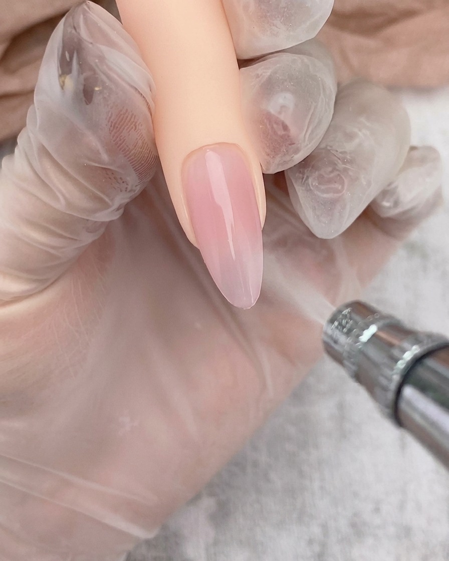 An airbrush tool applying a soft white gradient to the tip of a clear gel-coated nail.