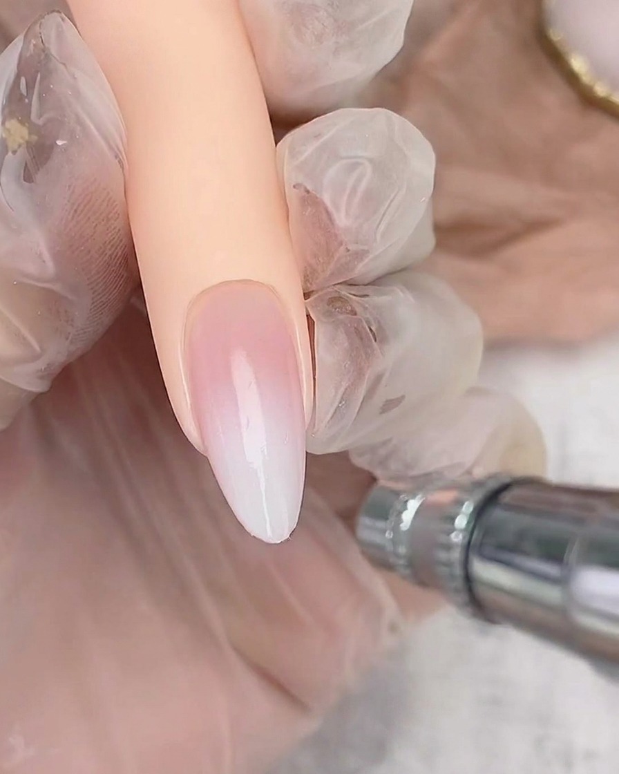 An airbrush tool lightly spraying white polish onto the free edge of an almond-shaped nail to build an ombre gradient.