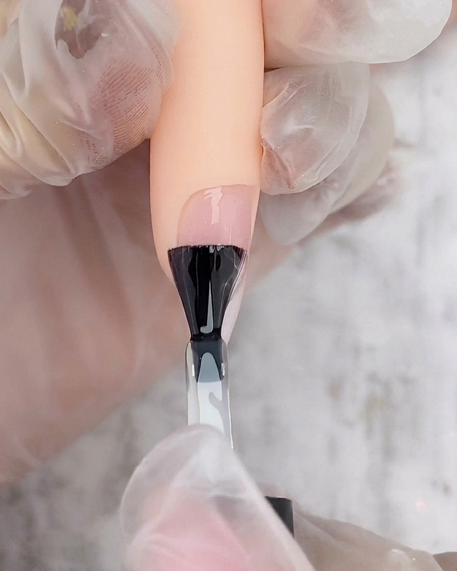 A black nail polish brush applying a layer of clear gel top coat over a white and pink ombre nail.