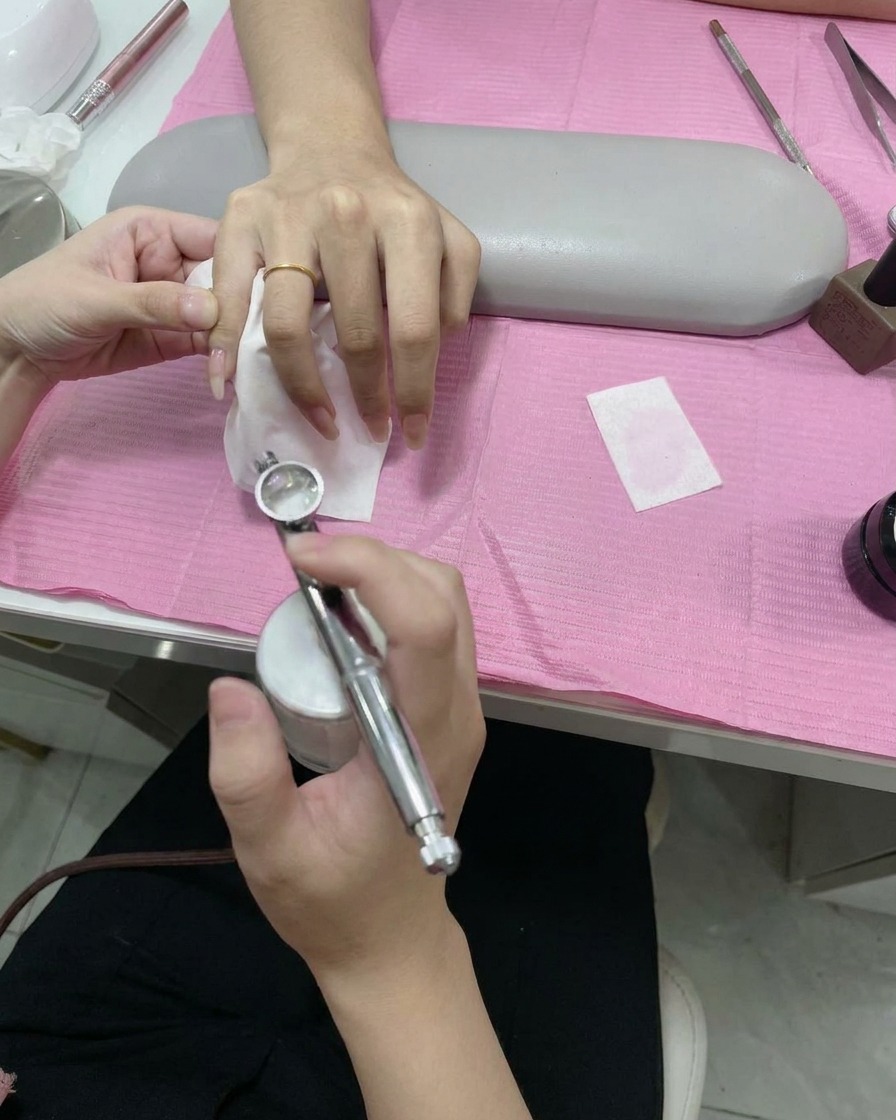 A hand holding an airbrush tool, spraying a fine mist of white paint onto the tip of a nail over a stencil.