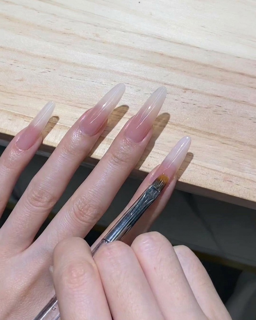 A fine-toothed ombre brush gently pulling wet milky white gel downwards on a clear stiletto nail extension.