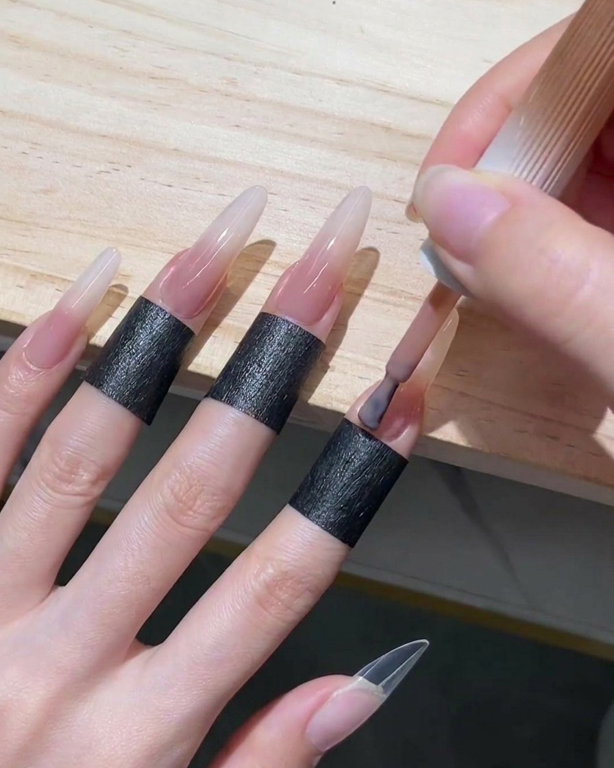 Applying a sheer nude-pink gel polish to the cuticle area and bottom half of a long stiletto nail.
