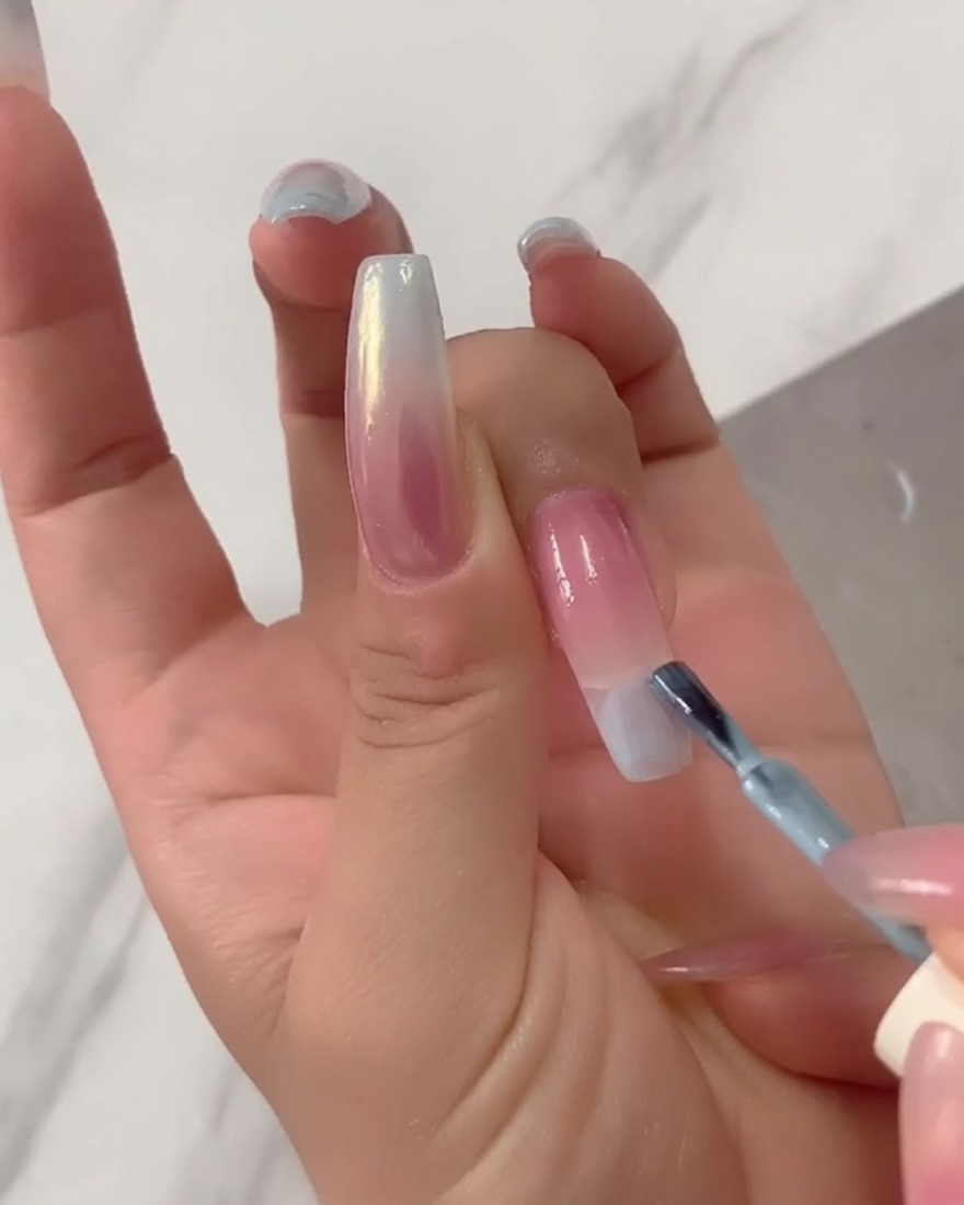 A close-up of a brush applying solid light blue gel polish directly to the tip of a long nail extension.