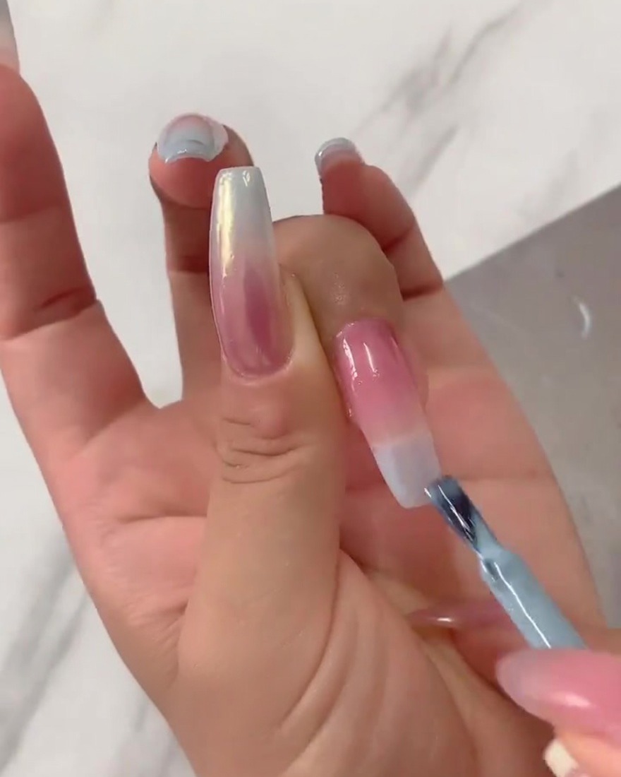 A close-up of a flat nail brush blending concentrated light blue gel polish downwards from the tip of a long fingernail over a sheer pinkish-nude base.