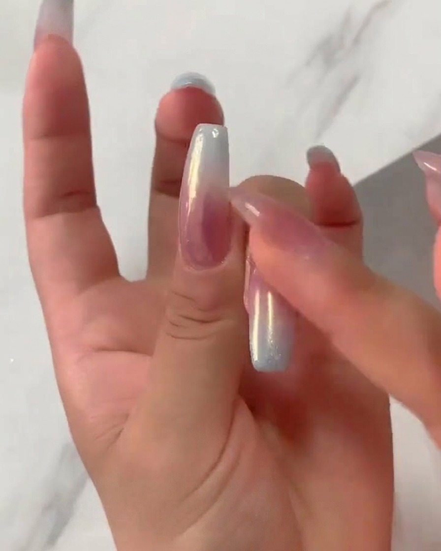 A fingertip firmly rubbing iridescent aurora chrome powder over a cured pink and blue gradient nail.