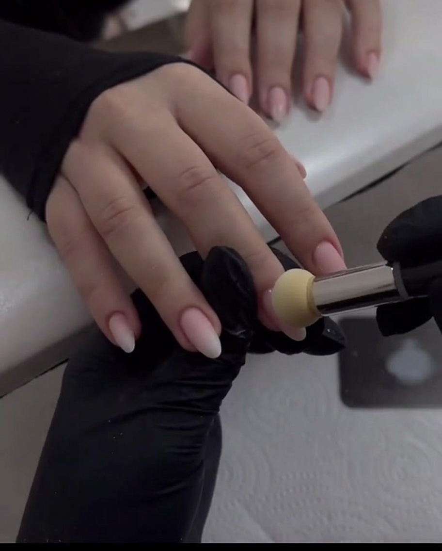 A gloved hand using a sponge applicator to buff pearl chrome powder downwards on a nude pink fingernail.