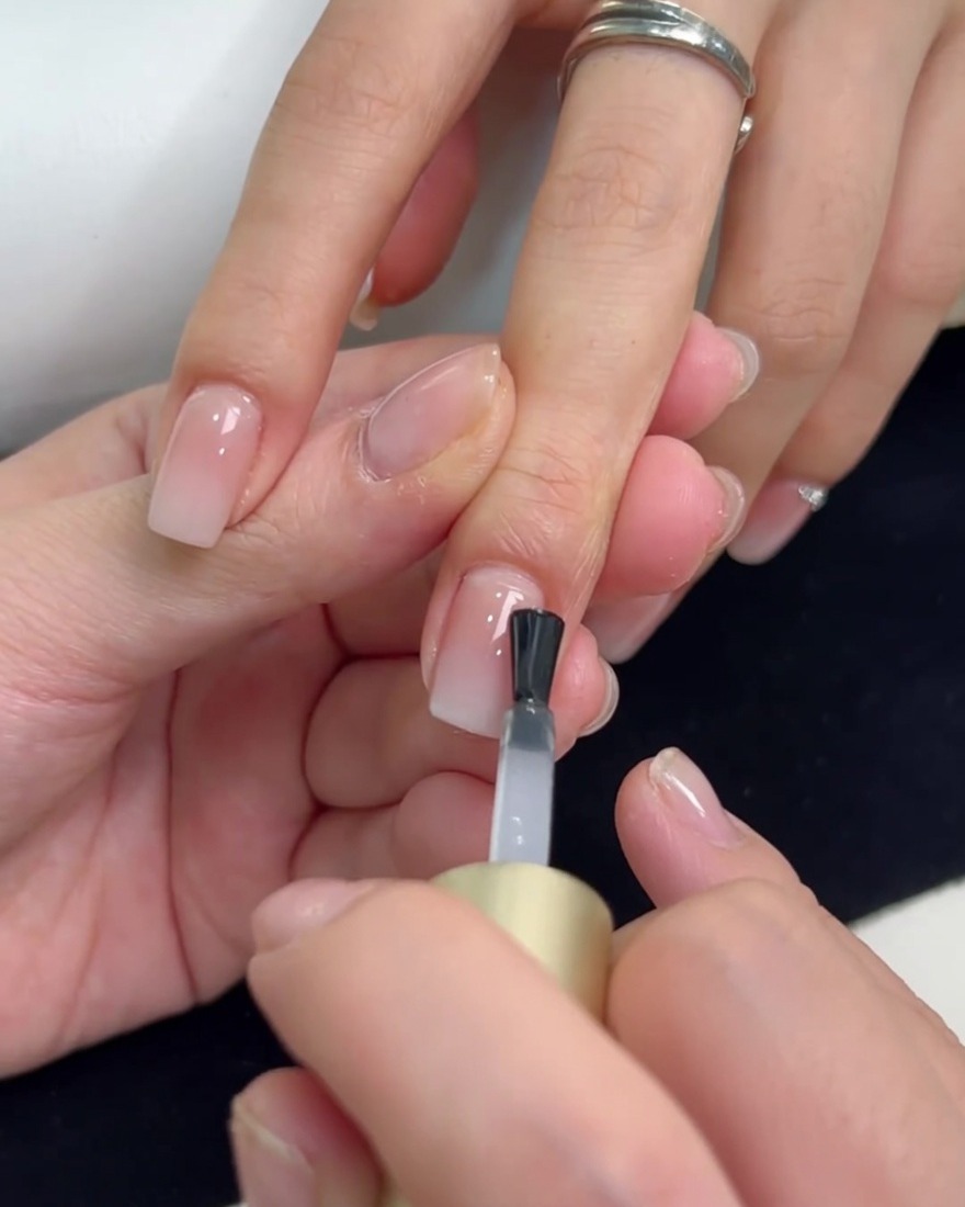 A brush applying a clear reinforcement top coat over a milky white gradient nail.