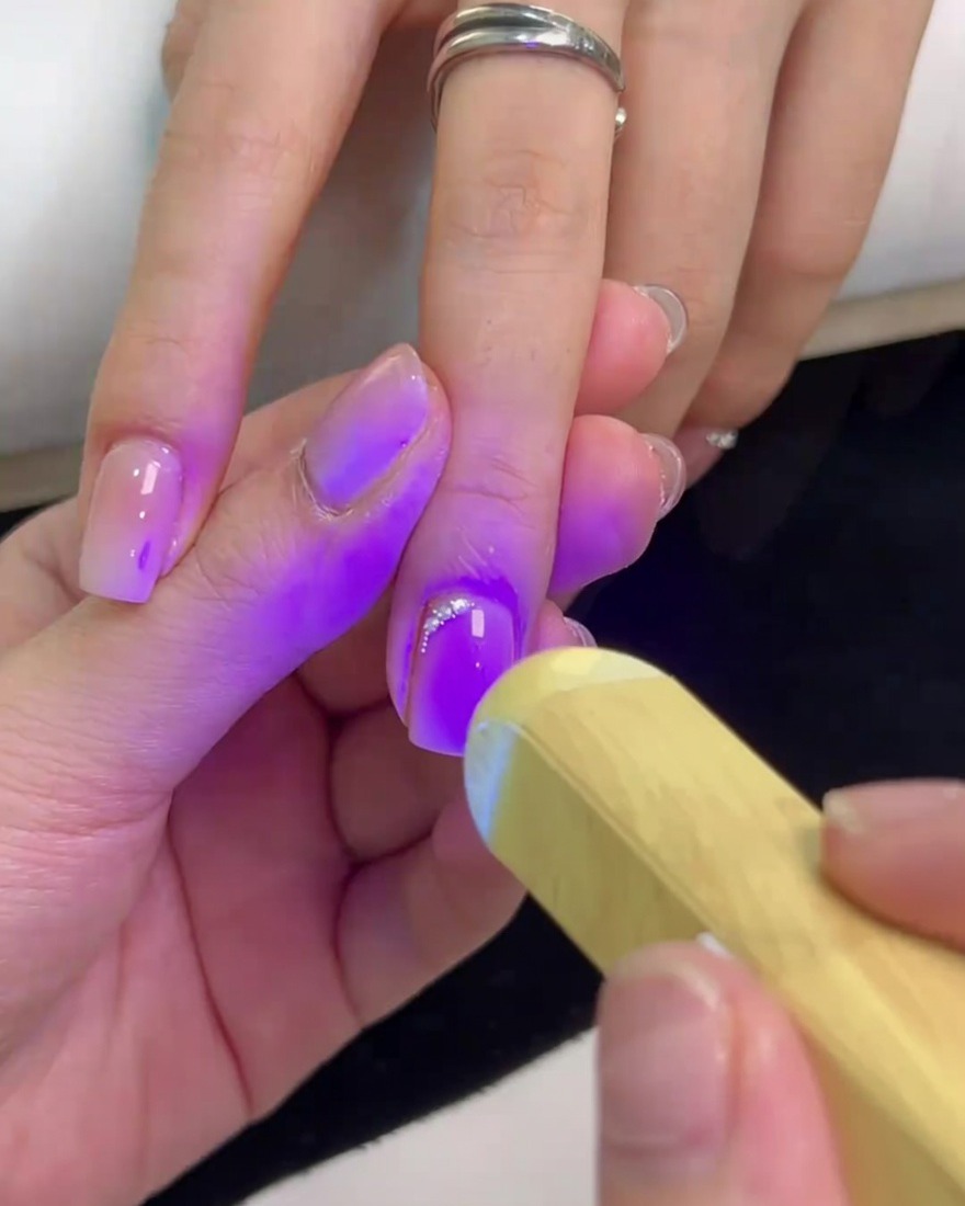 A handheld mini UV lamp shining a purple light onto a square nail to cure the rhinestone glue and secure the crystals.