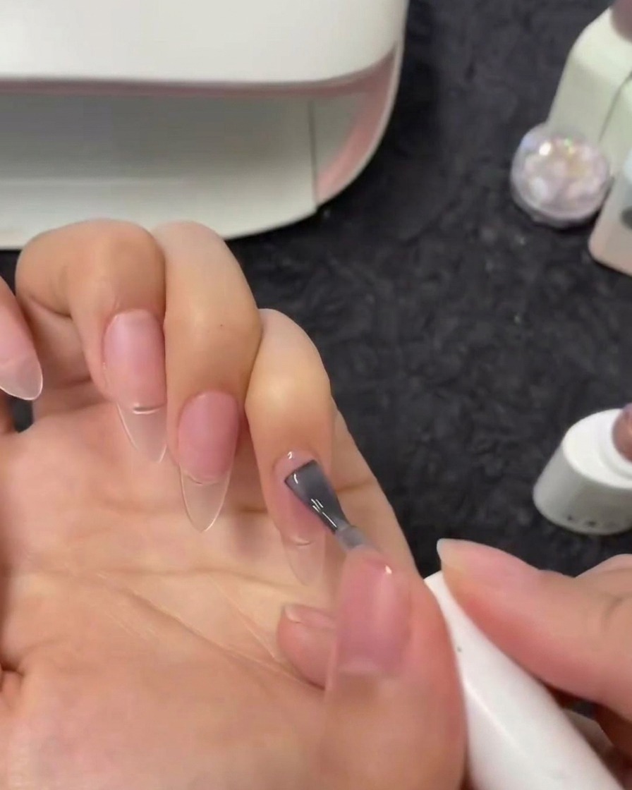 A close-up view of a brush applying a semi-sheer nude gel polish onto an index fingernail.