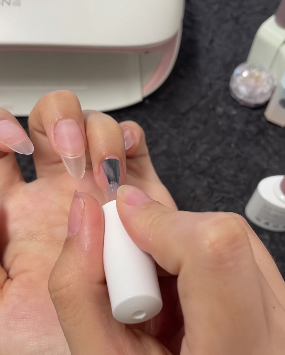 A close-up view of a brush applying nude gel polish to a middle fingernail.