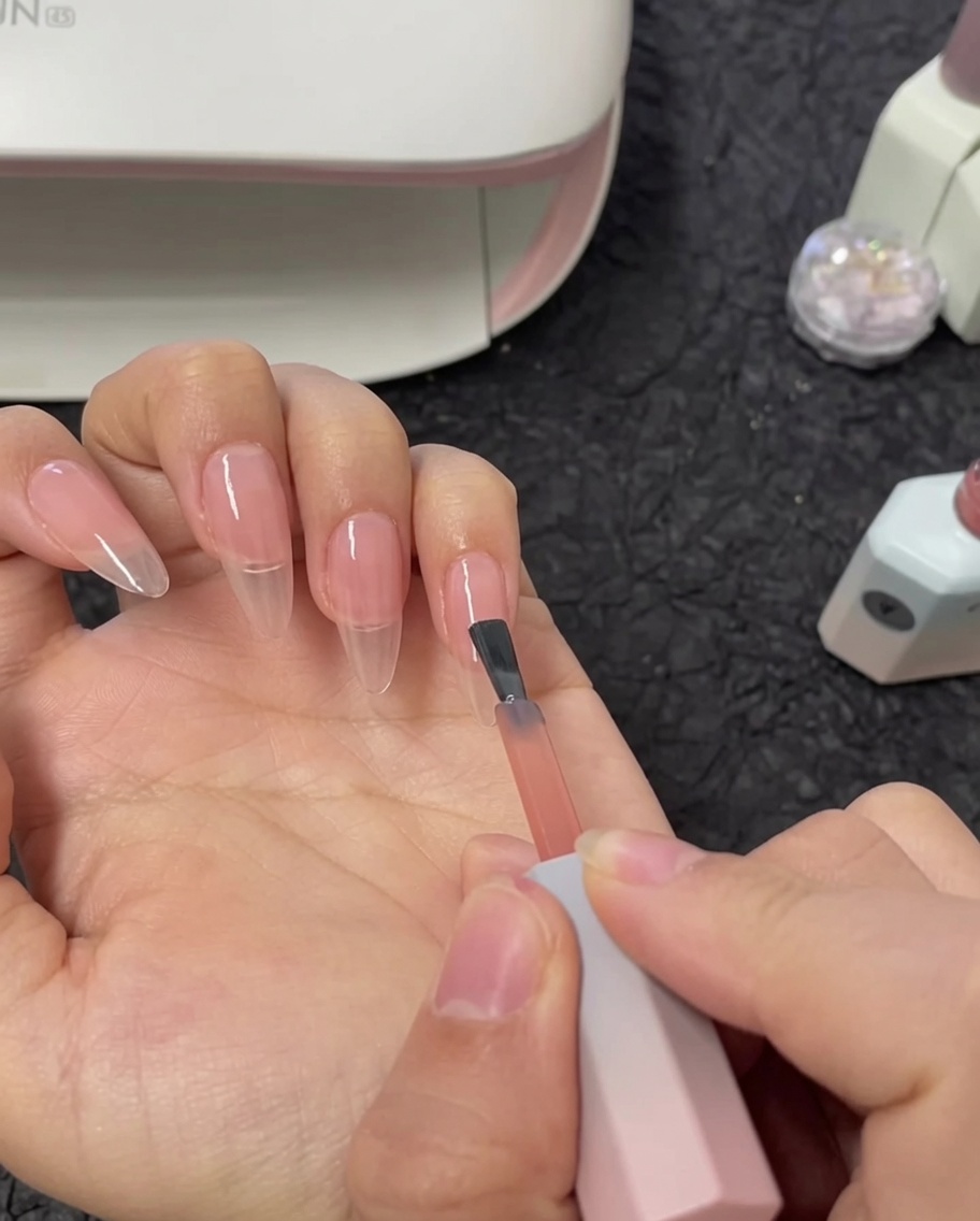 A brush applying a second layer of nude gel polish to an index fingernail.