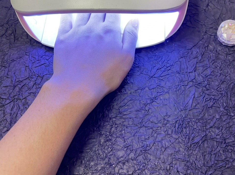 A hand resting inside a glowing UV/LED nail lamp to cure gel polish, sitting on a textured purple surface.