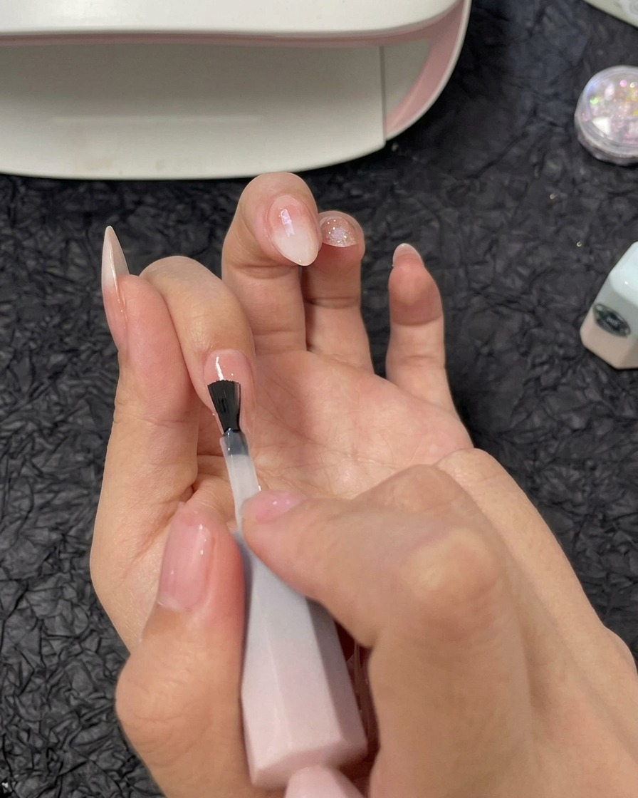 A hand applying a layer of clear builder gel over the ring finger nail using a gel polish brush.