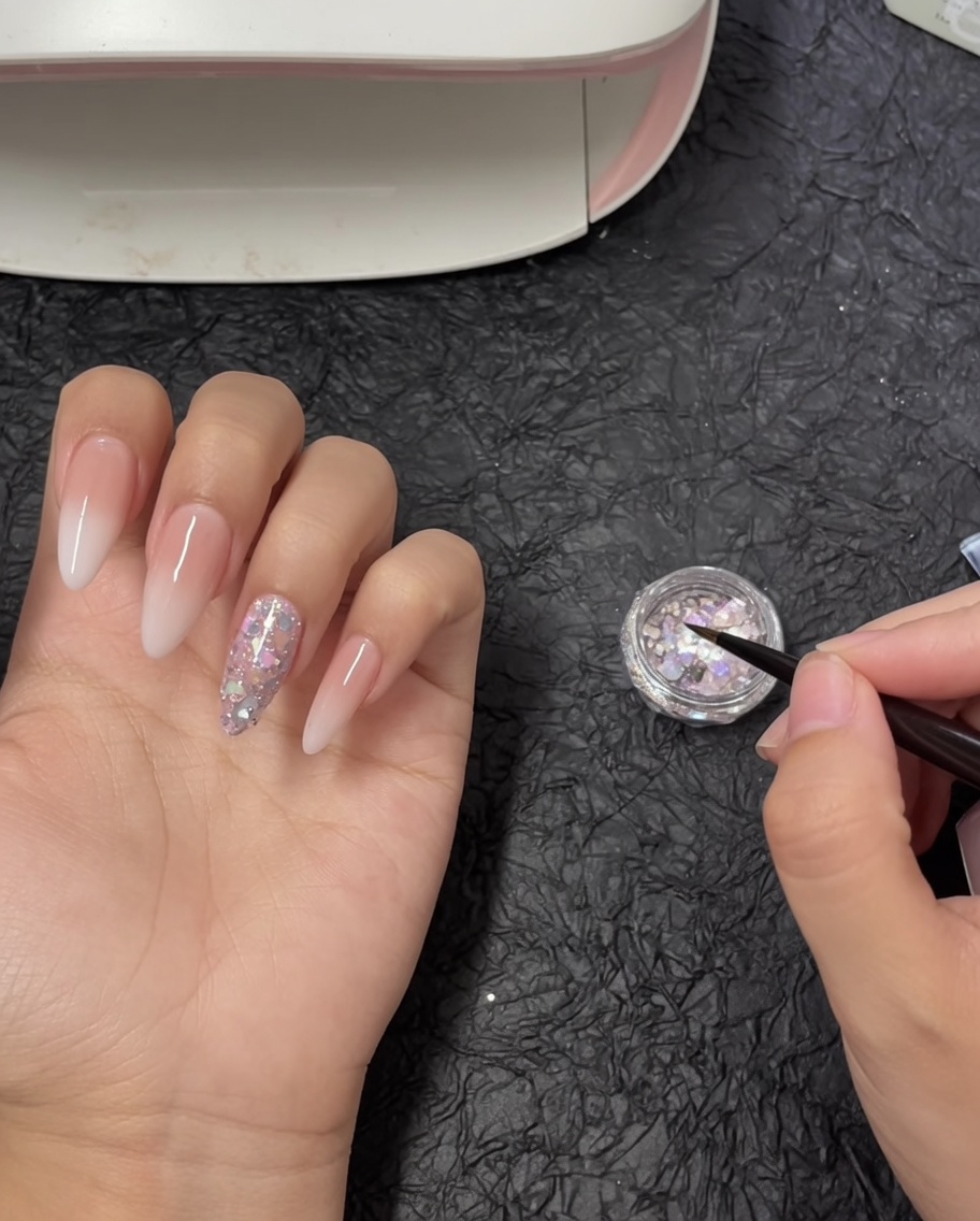 A detail nail art brush dipping into a small pot filled with iridescent chunky glitter flakes.