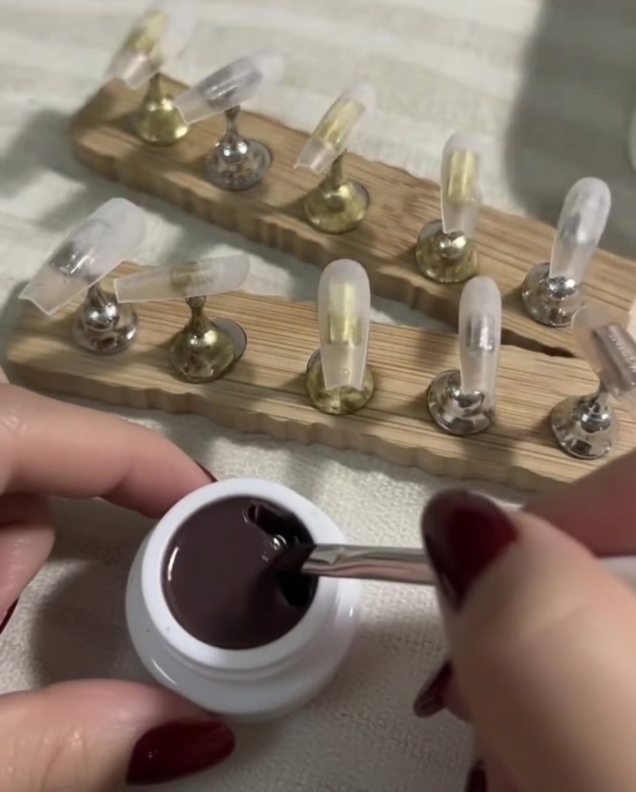 A flat gel brush dipping into a small jar containing a deep nude-toned gel polish.