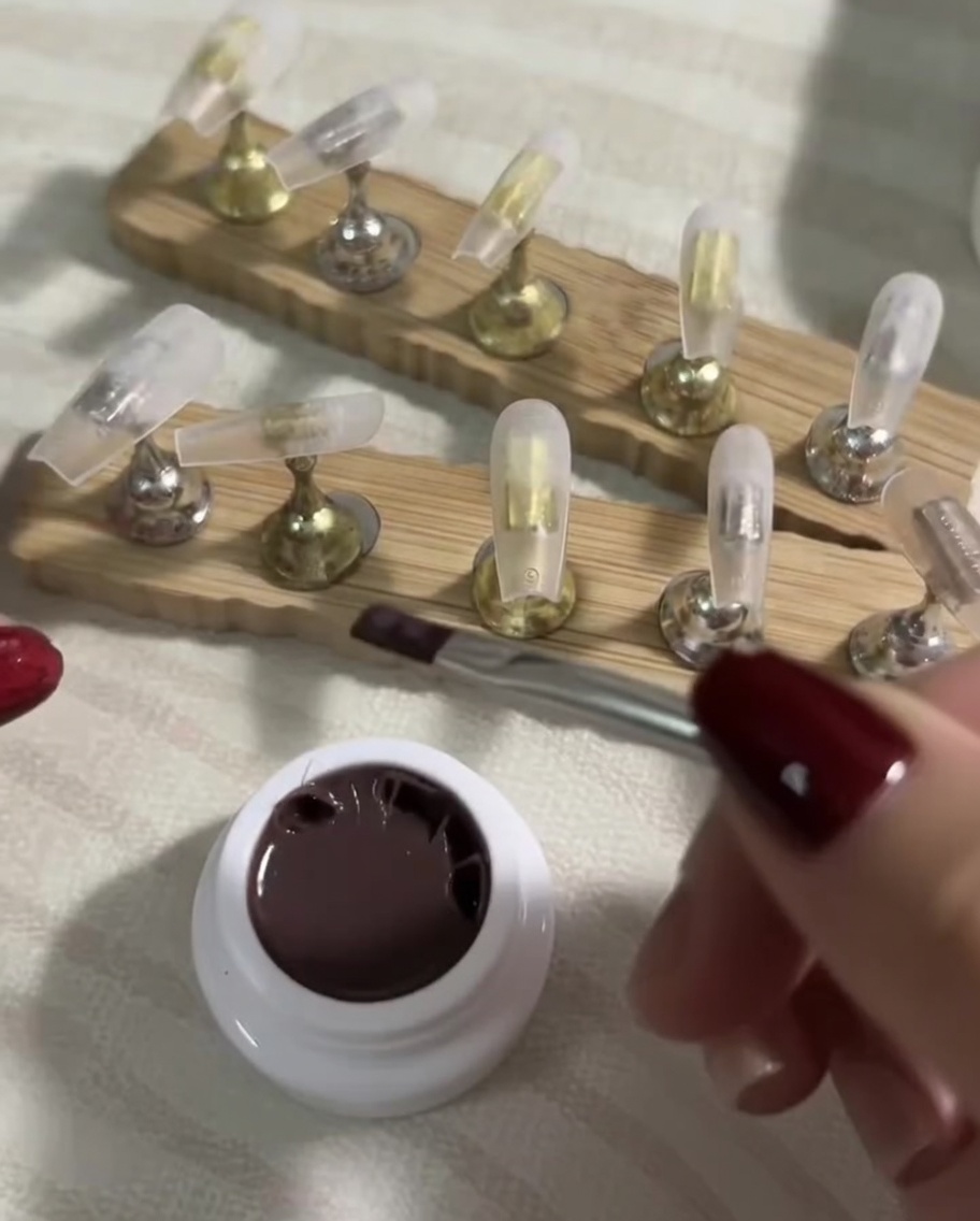 A flat brush smoothly applying a sheer coat of dark nude gel polish down a clear press-on nail tip.