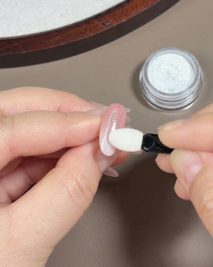 A hand using a small white eyeshadow sponge applicator to rub iridescent aurora powder onto a light pink gradient nail.