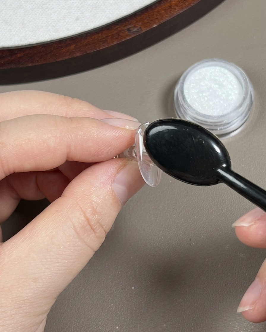 A black oval dusting brush sweeping over a polished nail to remove loose translucent chrome powder.