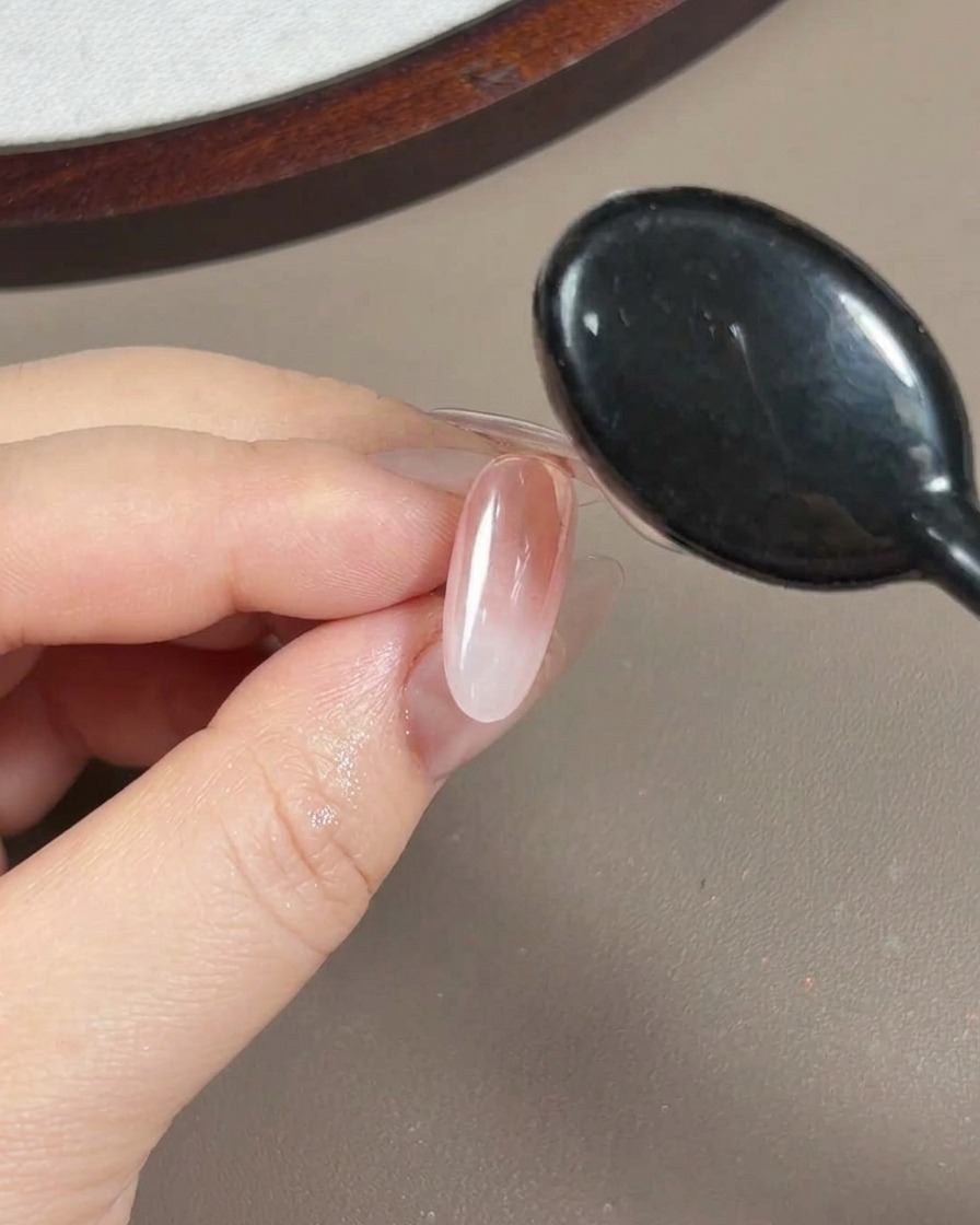 A close-up view of a brush applying a clear, viscous reinforcement gel over a nail that has been coated with aurora powder.
