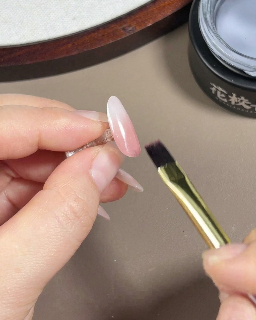 A gel nail brush applying a clear, glossy top coat onto a finished pink and white gradient nail with an aurora finish.