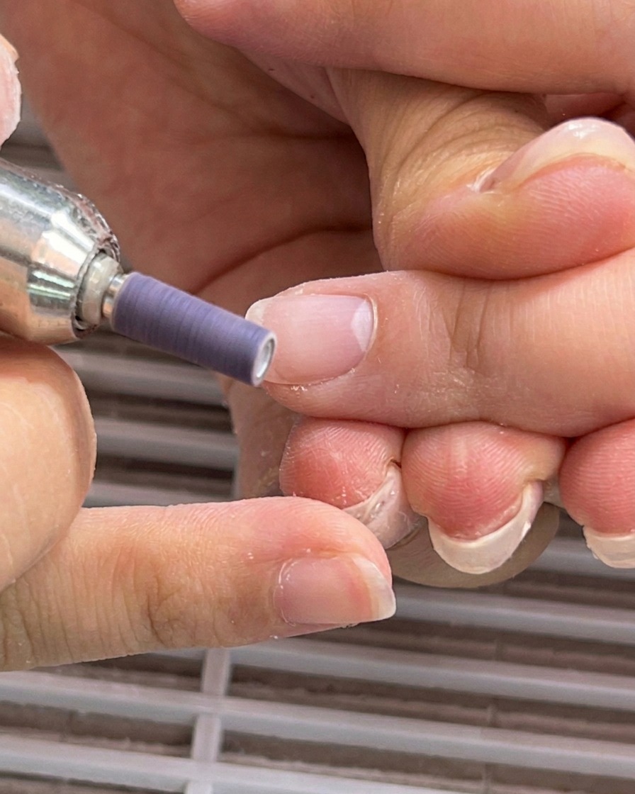 An electric file with a purple sanding band smoothing the nail plate near the cuticle fold.