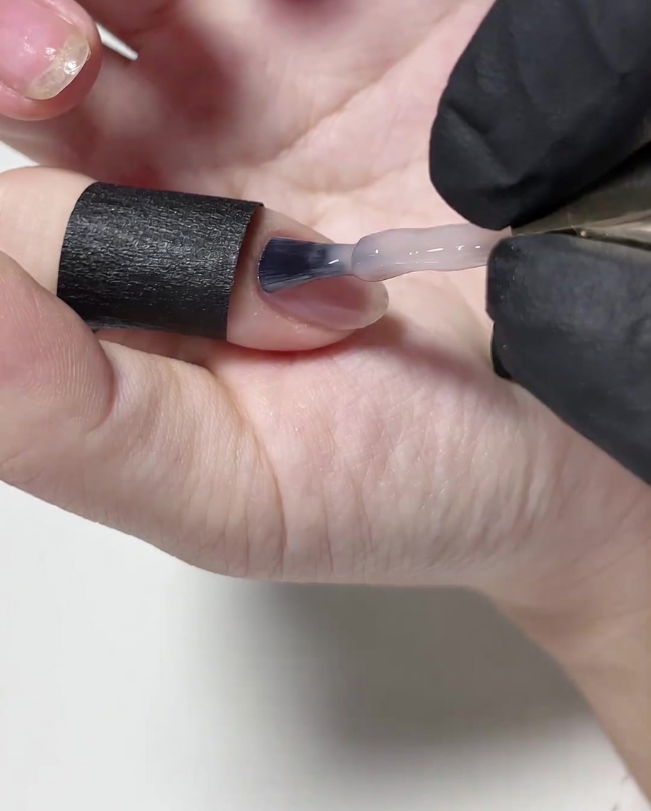 A brush applying a thin layer of nude gel polish onto the surface of the nail.