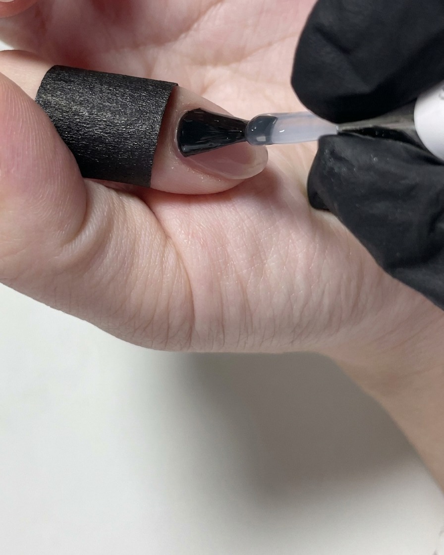 A nail technician applying clear matte top coat over a nude gel nail using a black bottle brush.