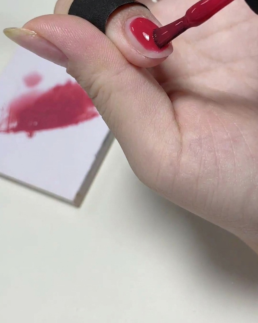 A gel brush applying a sheer layer of pink polish to the top half of a fingernail, with a mixing palette visible in the background.