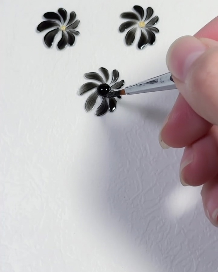 A dotting tool placing a precise yellow dot into the center of a black daisy design.