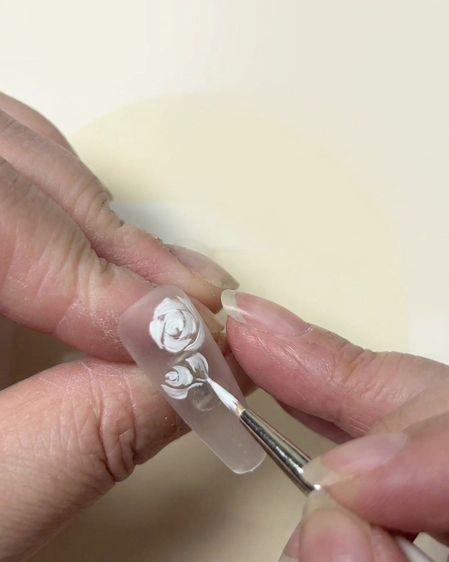 A painterly close-up of a white rose design being refined with a fine-tipped brush on a clear nail.