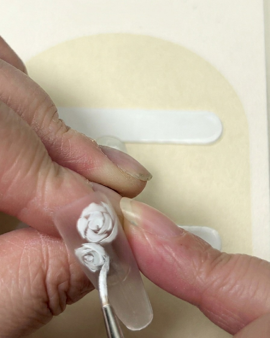 A close-up of a five-petaled flower being painted in white gel next to an established rose design.