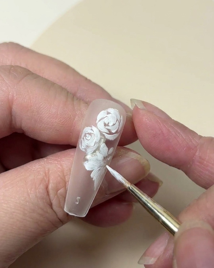 A close-up of a nail tip showing a white painted rose being complemented by a small five-petaled flower and leaf details.