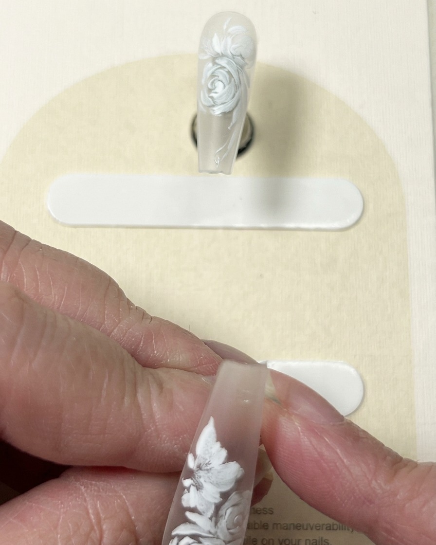 A close-up view of a brush adding fine white detail lines to the center and petals of a painted floral design on a nail.