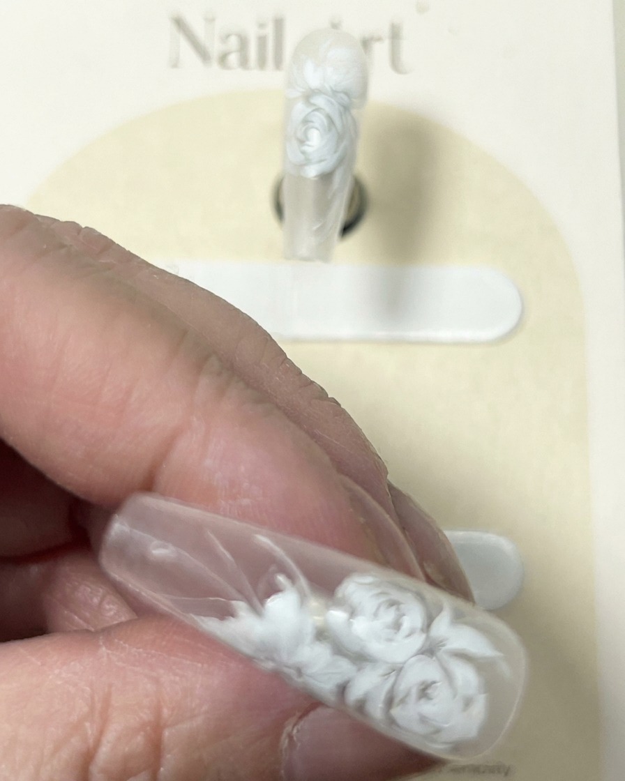 A close-up of a nail with a detailed white rose design, showing the brush adding final delicate white highlights.