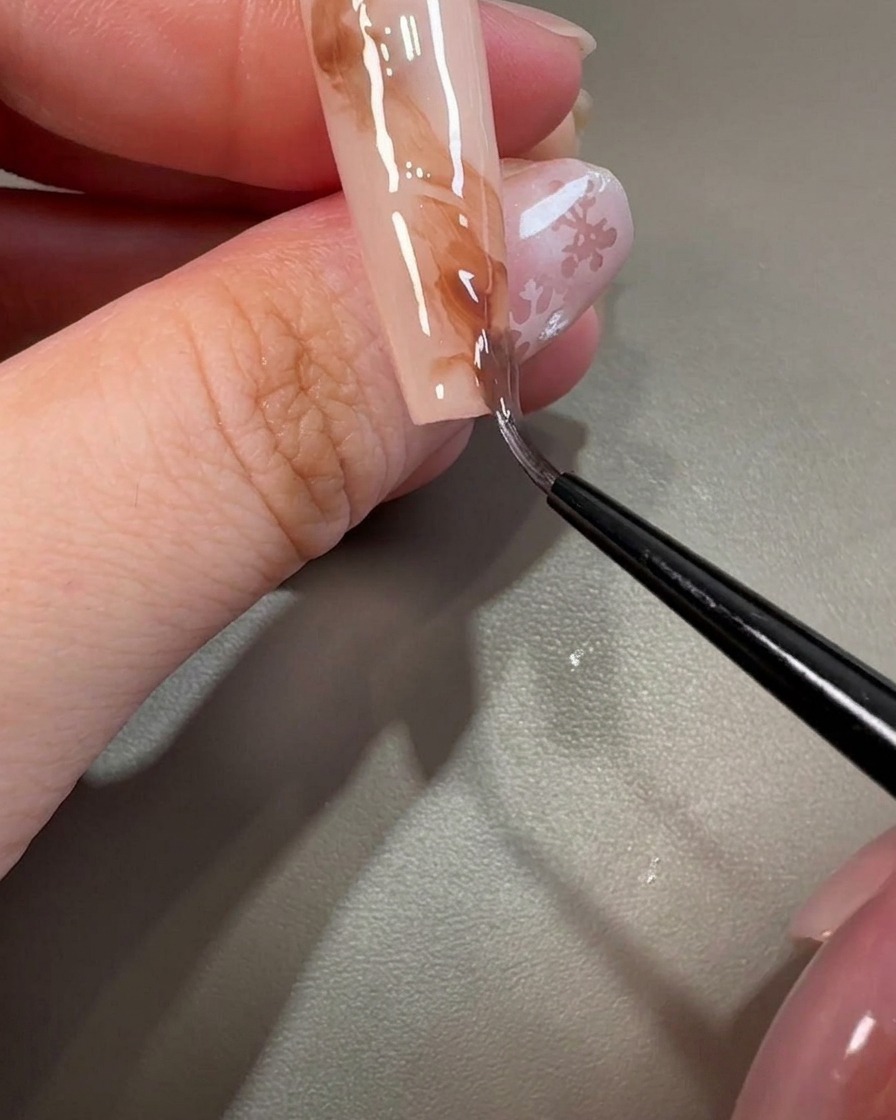 A liner brush pulling through brown gel to create swirling, marbled textures on the nail.