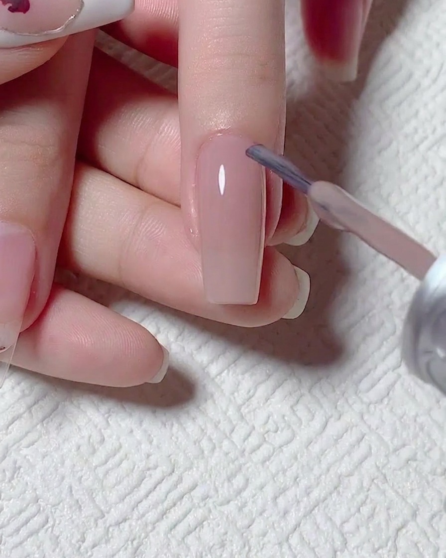 A close-up of a brush applying a sheer, nude gel polish to a prepared nail surface.
