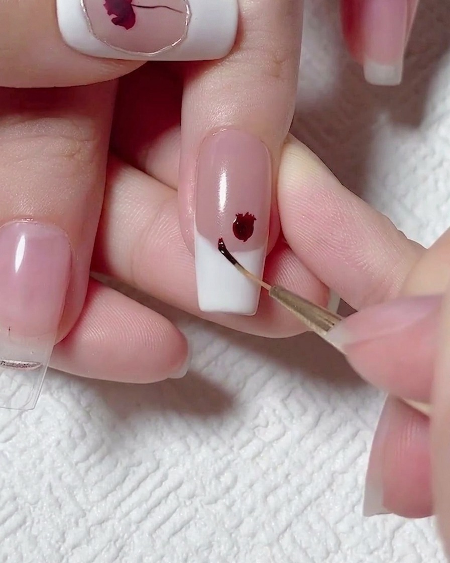 A close-up of a liner brush applying a tiny dot of dark red gel polish beside a larger painted red flower on a French tip nail.
