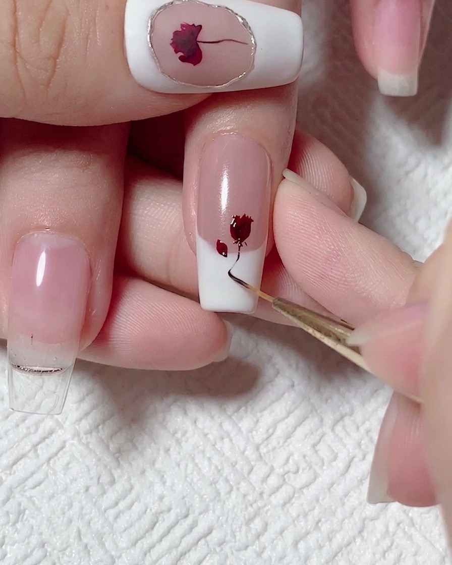 A liner brush pulling a thin, curving line of black-brown gel downwards from a painted red rose on a fingernail.