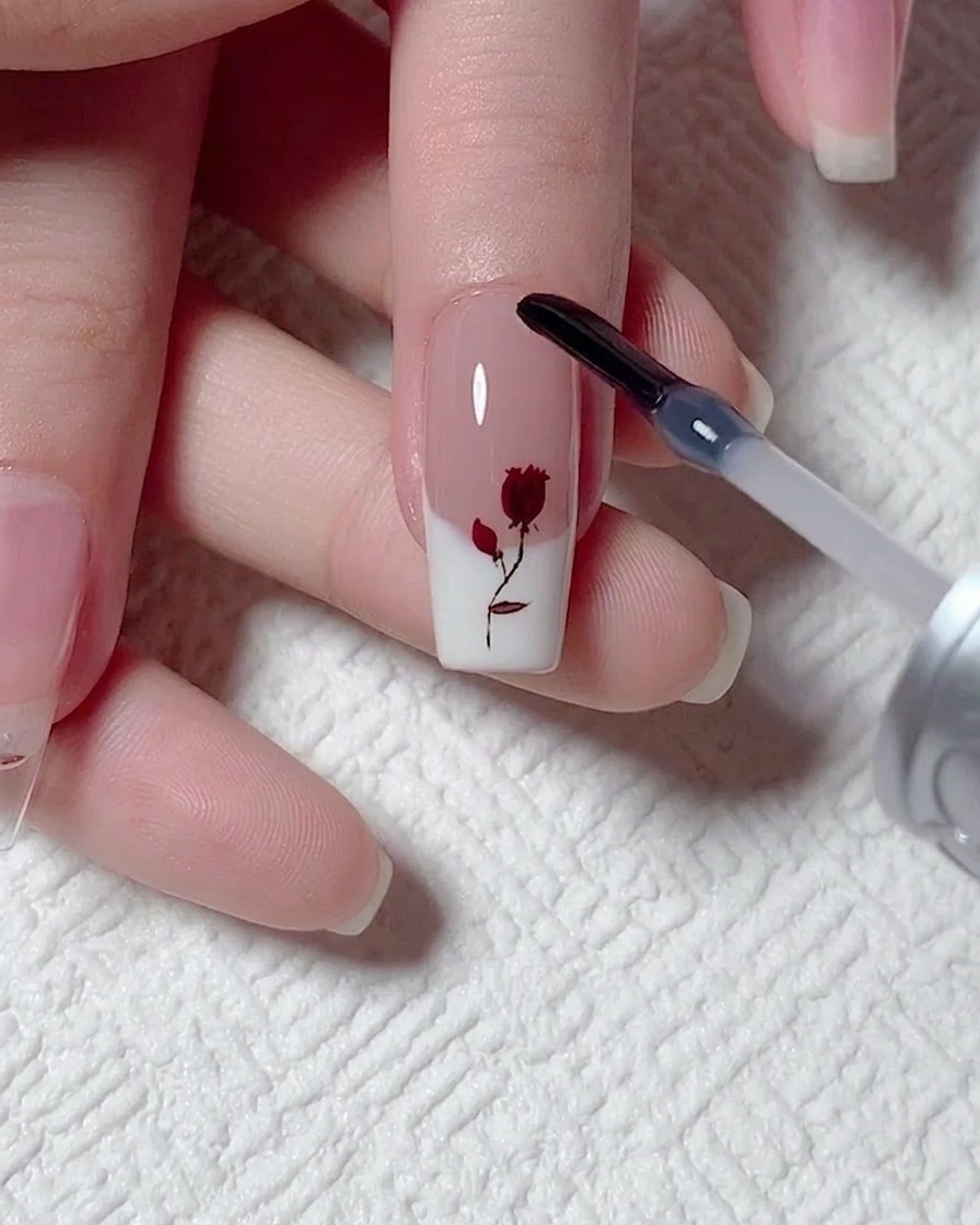 A gel brush from a bottle applying a smooth, clear layer of reinforcement gel over a painted rose nail design.