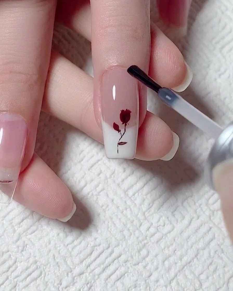Applying a layer of matte top coat with a bottle brush over a nail featuring a red rose and white French tip design.