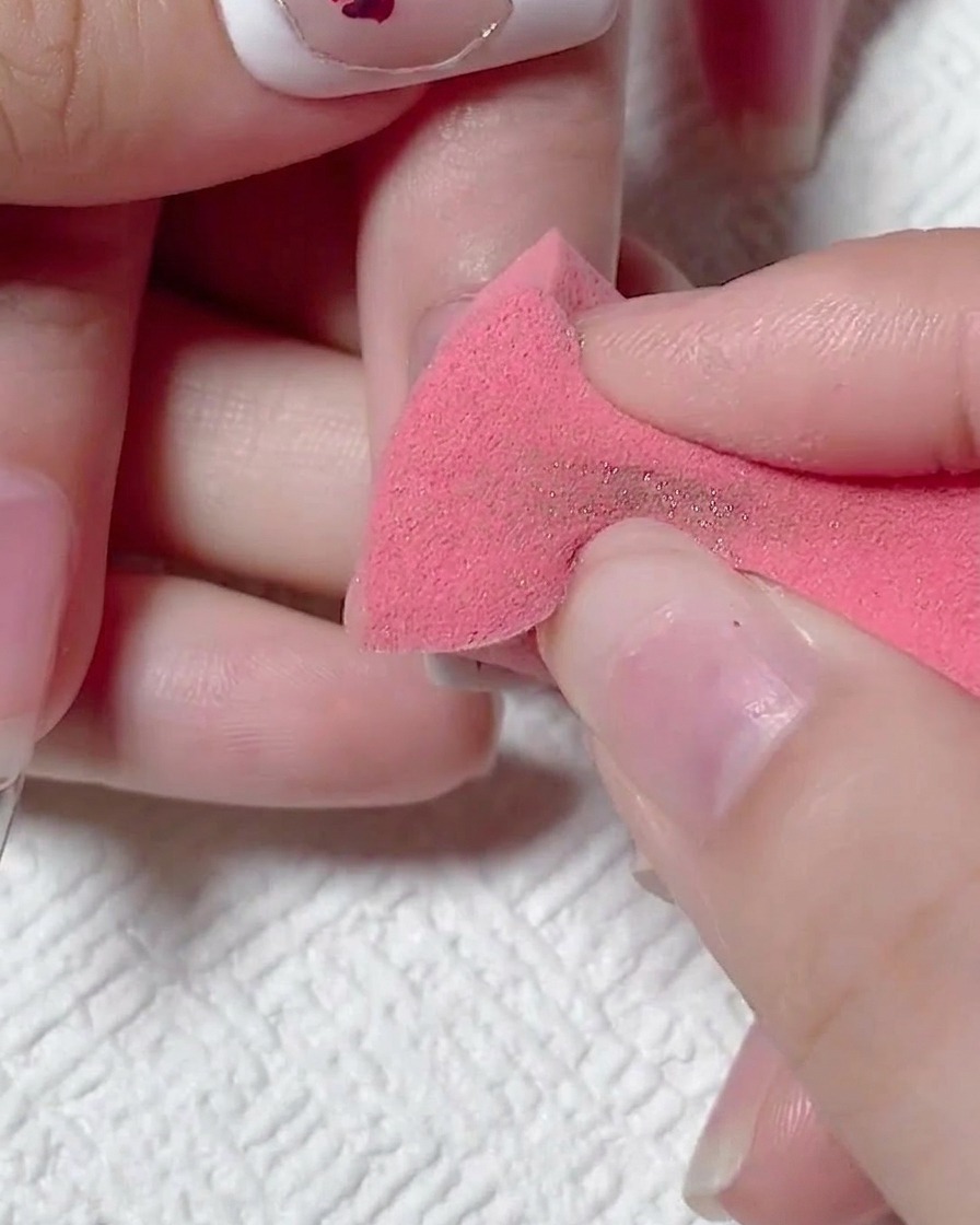 A hand using a dry pink makeup sponge to wipe away excess gold chrome powder from a matte nail.