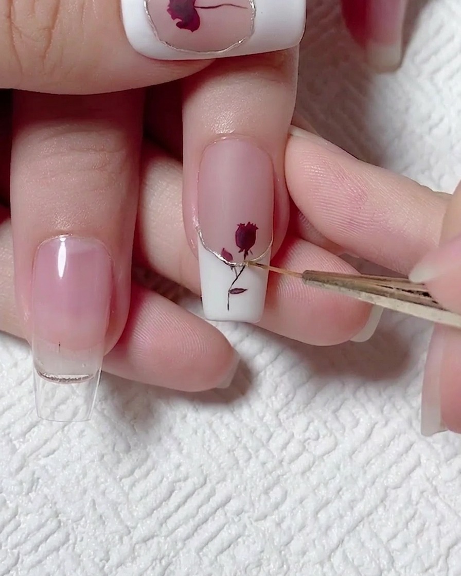 A close-up view showing a fine liner brush applying a precise line of top coat over a metallic champagne-colored accent on a white French-tip nail.