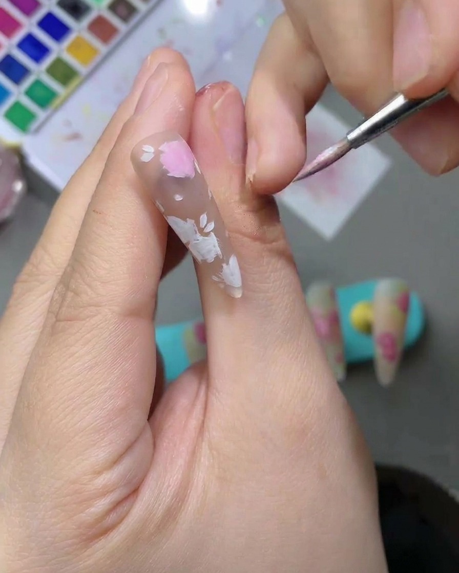 A close-up of a nail with delicate light pink watercolor petal shapes being filled in using a fine-tipped nail art brush.